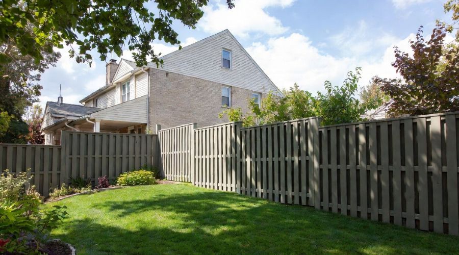 A fenced-in backyard with a green lawn, garden beds, and a large multi-story brick house under a partly cloudy sky.