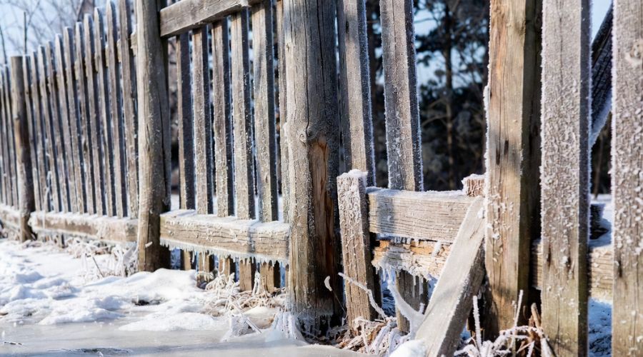 A weathered wooden fence covered in frost and ice, standing in a snowy landscape during a sunny winter day.