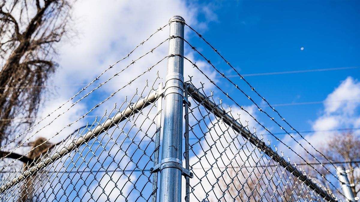A chain-link fence corner with barbed wire extending across the top, set against a blue sky with scattered white clouds.