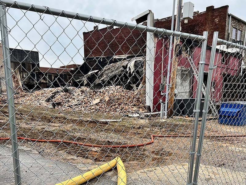 A chain-link fence in the foreground overlooks a pile of rubble from a building that has partially collapsed.