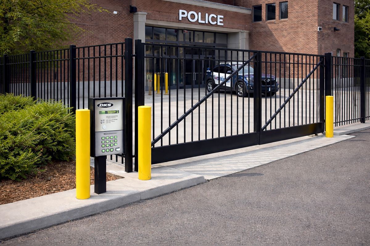 A black metal security gate blocks the entrance to a brick police building, with two yellow protective bollards in front.
