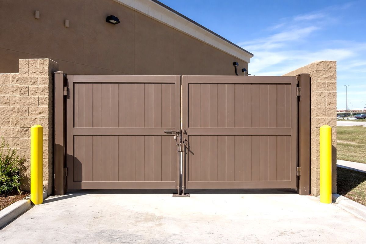 Double-door brown gate set between tan brick walls with yellow safety bollards on a concrete pad.