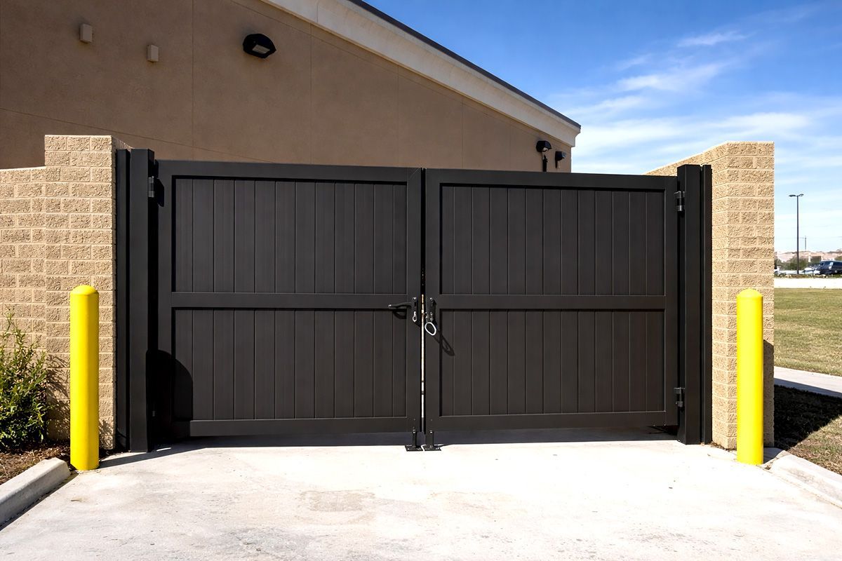 A pair of dark, vertical-slatted metal gates between brick pillars, flanked by two bright yellow safety bollards.