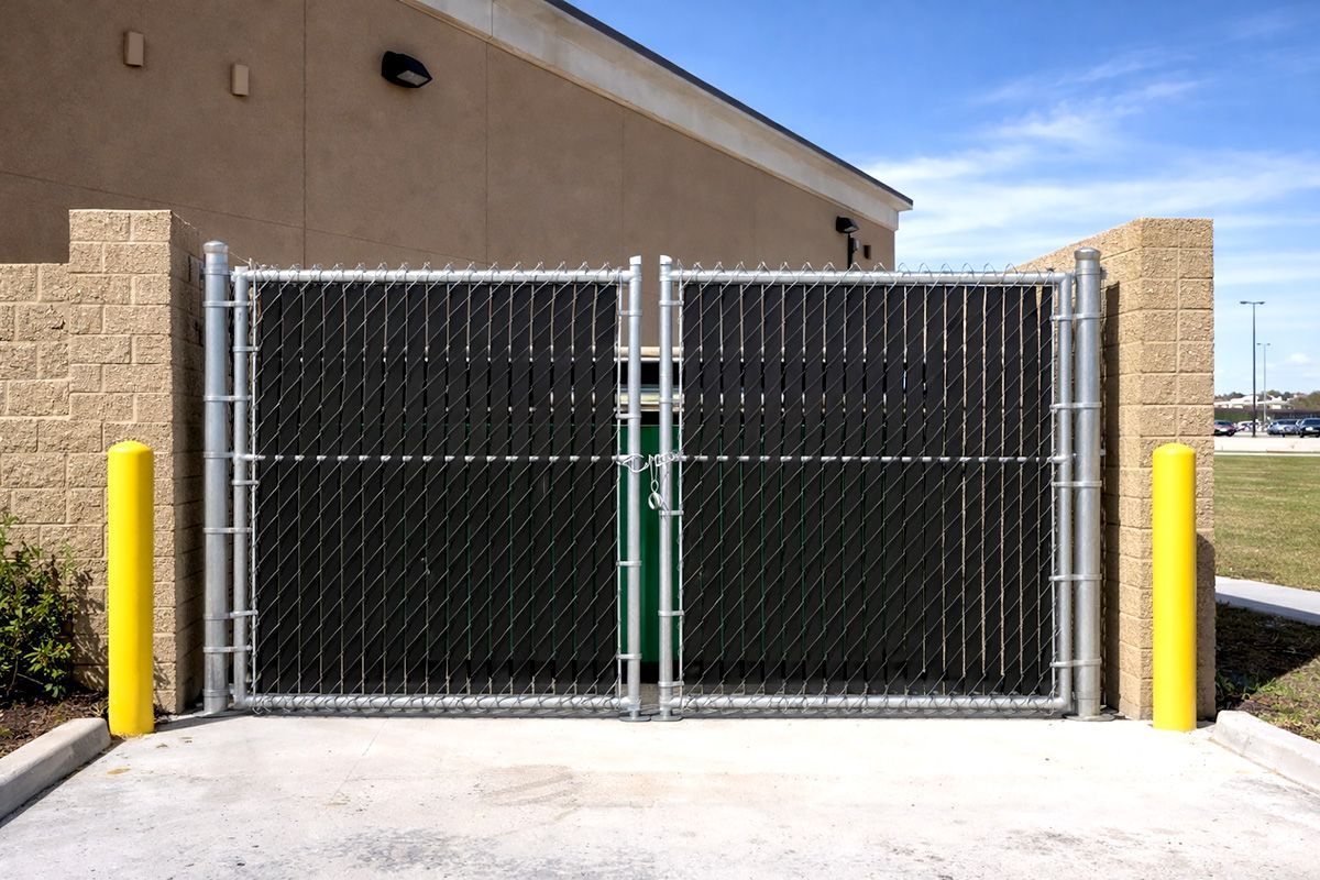 Double gate with black privacy slats set between two brick walls and yellow safety bollards.