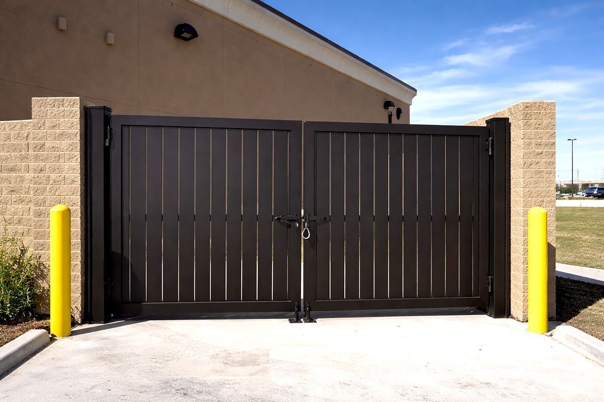 A closed, dark-brown metal double gate set between two tan brick walls, flanked by two yellow bollards on a concrete pad.