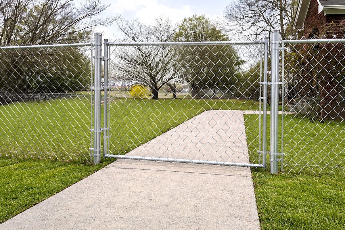 A silver metal chain-link gate and fence installed across a concrete sidewalk in a residential yard.
