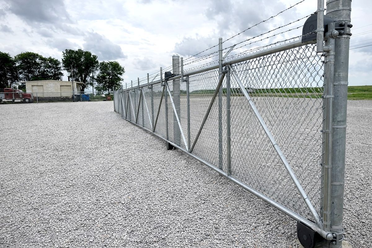 A galvanized steel chain-link gate with barbed wire topping, standing on a gravel lot under a cloudy sky.