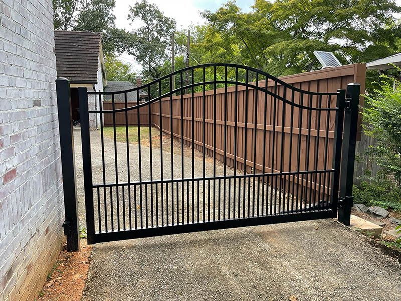 A black arched metal driveway gate installed between a brick building and a wooden privacy fence.