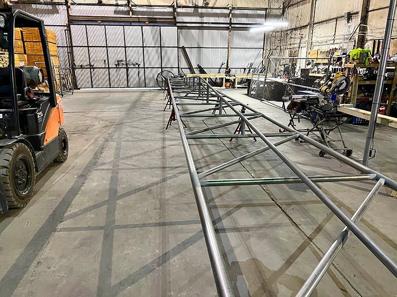 A long, narrow metal truss structure rests on stands inside a warehouse next to a forklift.