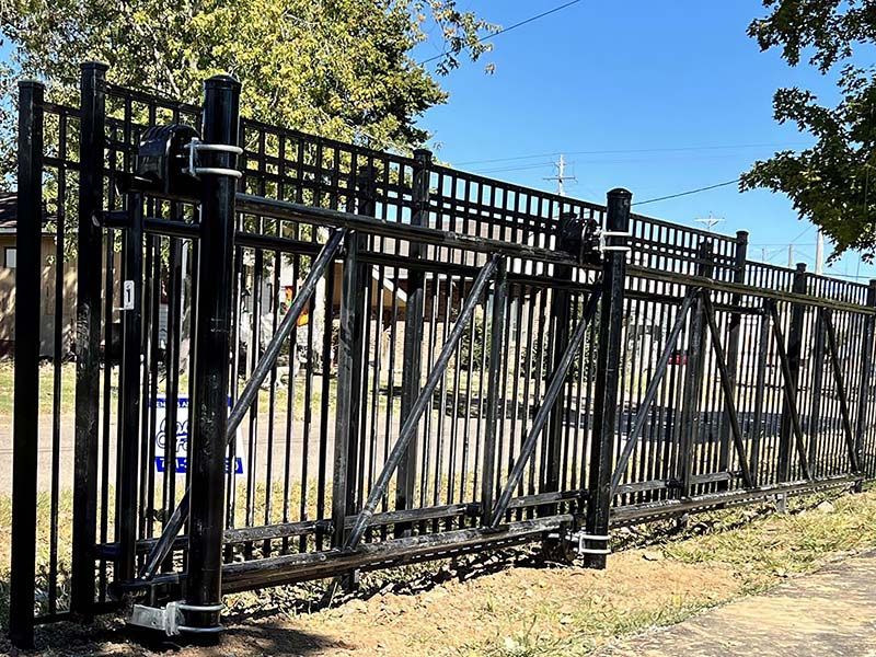 A black metal sliding gate with diagonal support beams, mounted on heavy poles, standing outdoors on a sunny day.