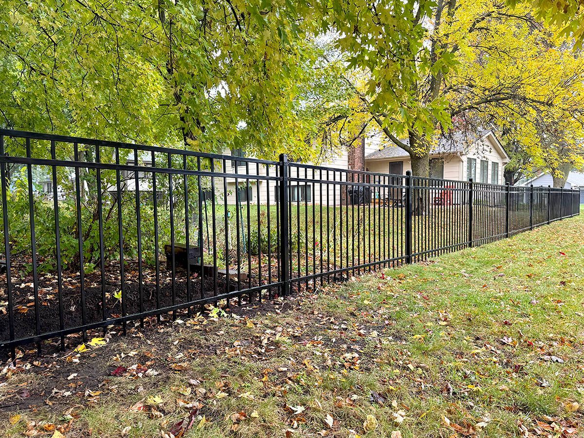 A black metal fence runs across a lawn covered in fallen leaves, with a house visible in the background among autumn trees.
