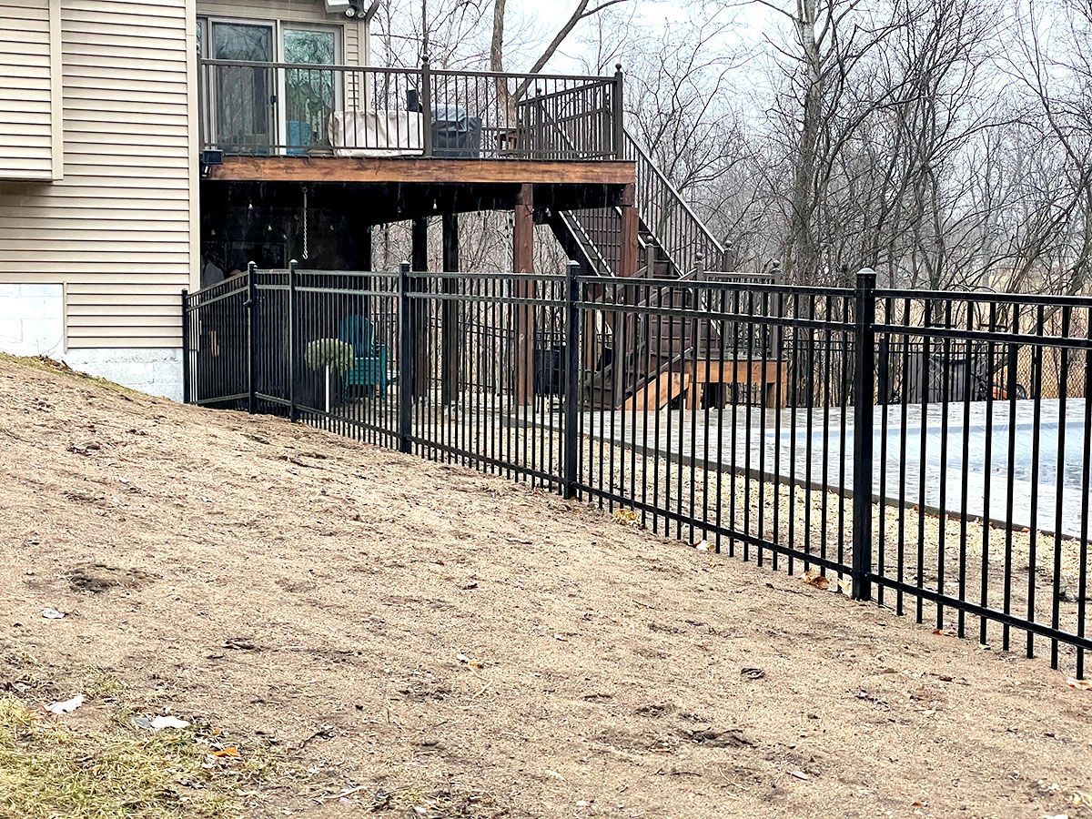 A black metal fence stands on a grassy slope leading toward a house deck and stairs in a backyard setting.