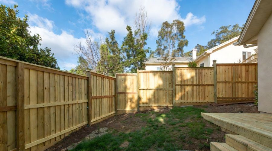 A wooden privacy fence surrounds a grassy backyard, with a wooden deck in the foreground and trees under a blue sky.