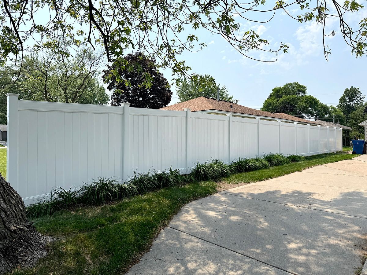 A long, white vinyl privacy fence runs alongside a concrete driveway on a sunny day with trees in the background.