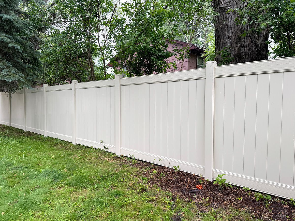 A long, light-colored vinyl privacy fence borders a grassy yard with trees in the background.