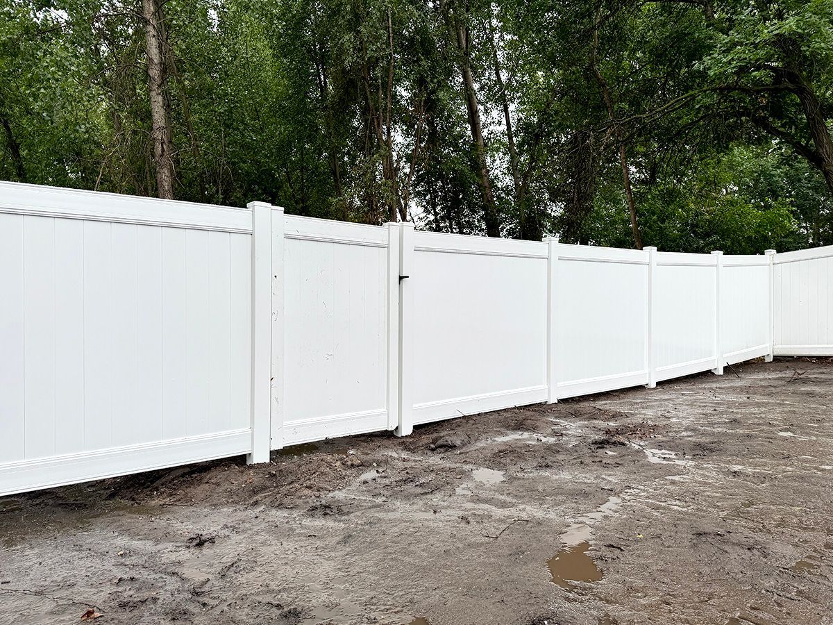 A newly installed white vinyl privacy fence set against a line of trees in an unfinished dirt yard.