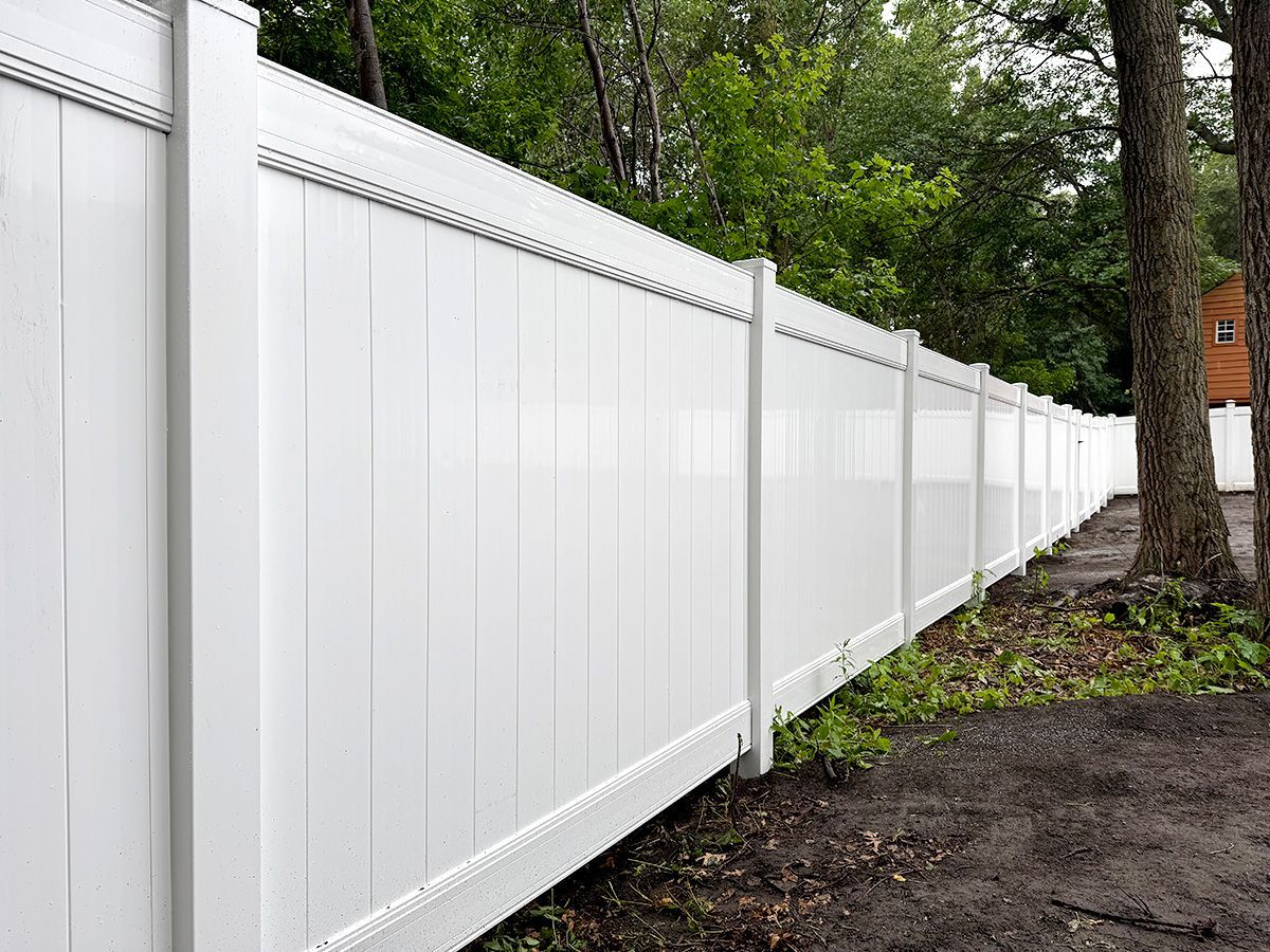 A long, white vinyl privacy fence stands in a wooded area, extending from the foreground into the background.