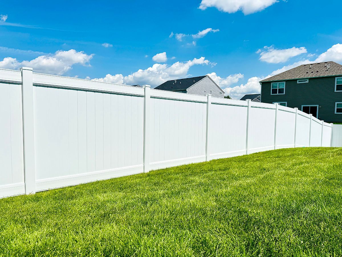 A long, white vinyl privacy fence stands on a grassy lawn under a blue, partly cloudy sky near a house.