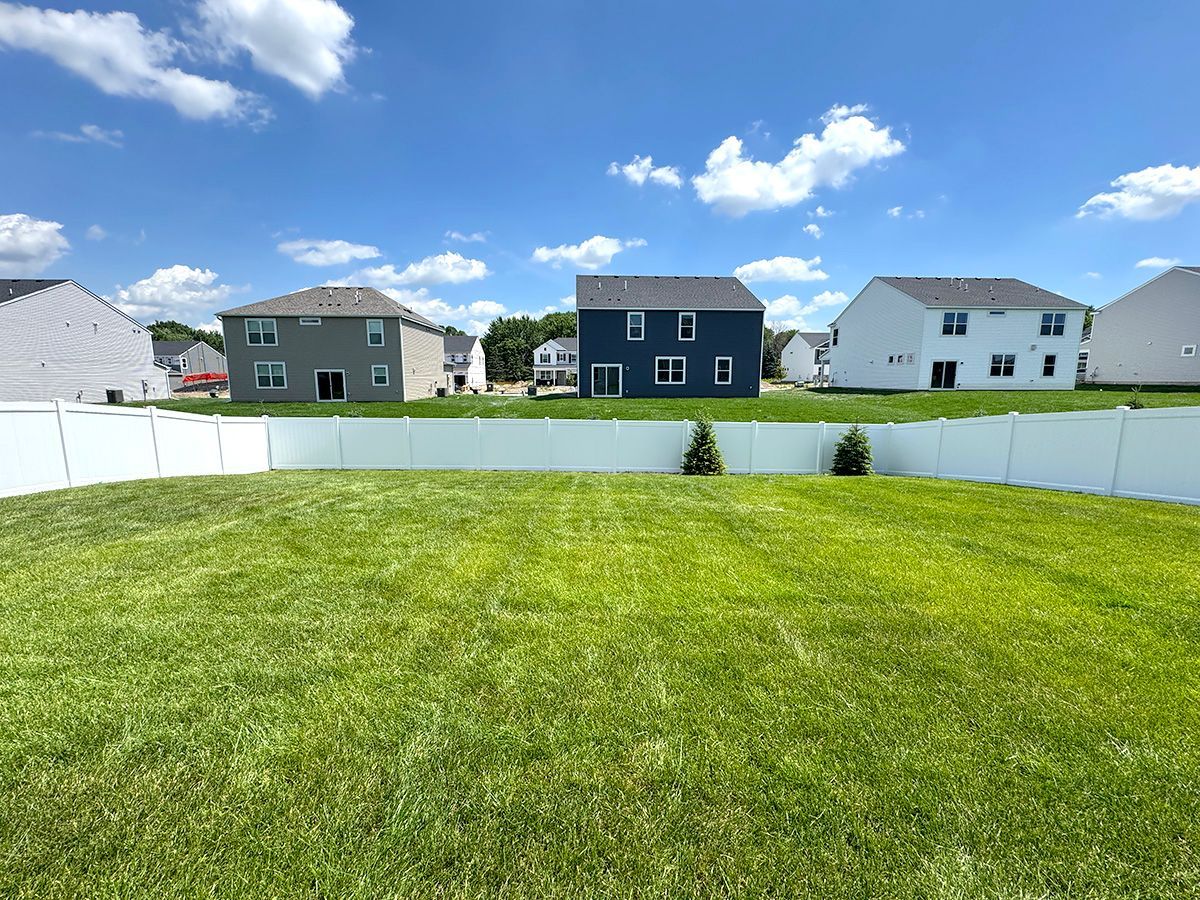 A lush green lawn fenced in white with several suburban houses in the background under a blue sky with white clouds.