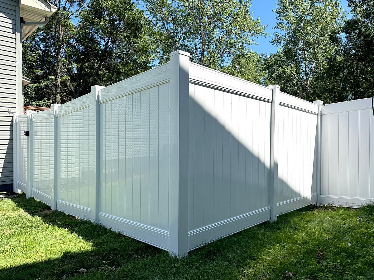 A white vinyl privacy fence stands in a grassy backyard next to the side of a house, set against a backdrop of trees.