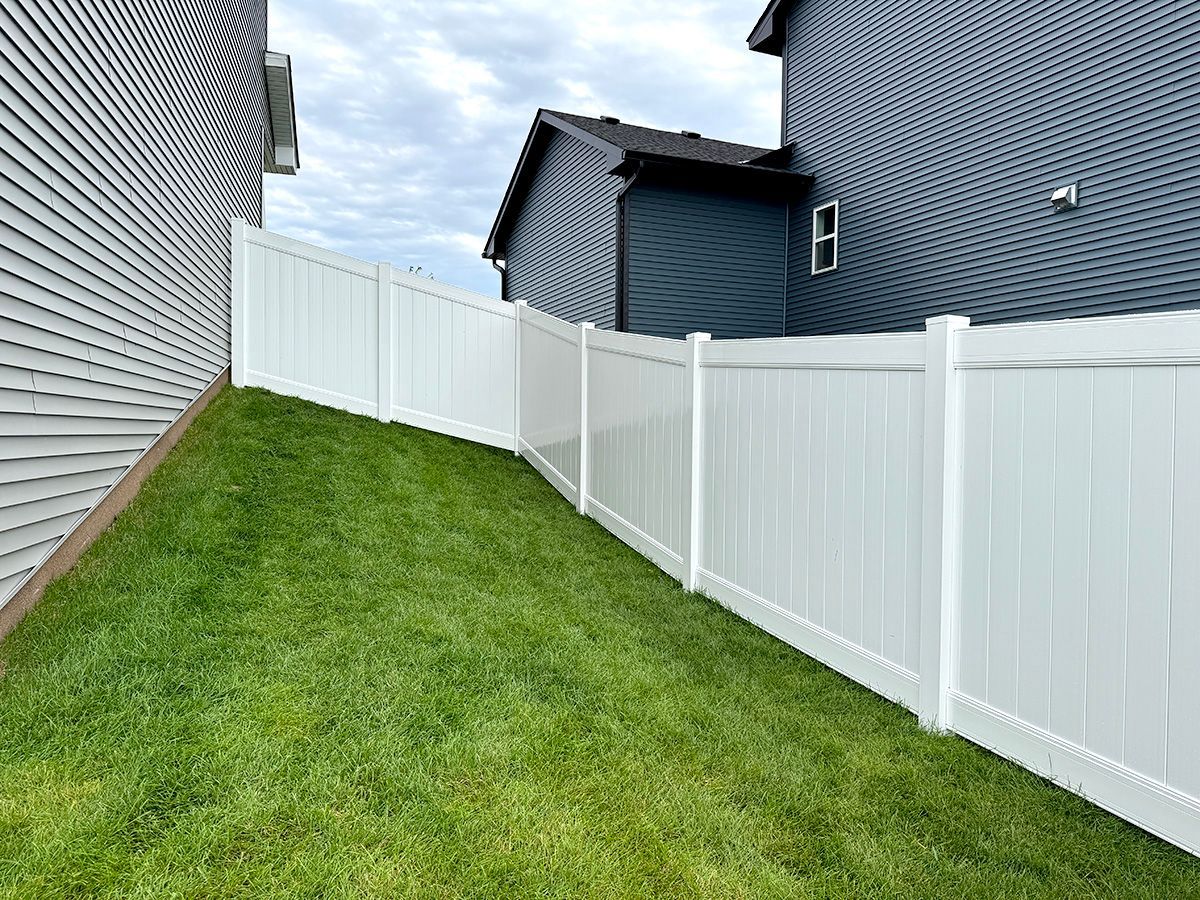 A white vinyl fence runs along a grassy slope between two houses with grey siding under a cloudy sky.