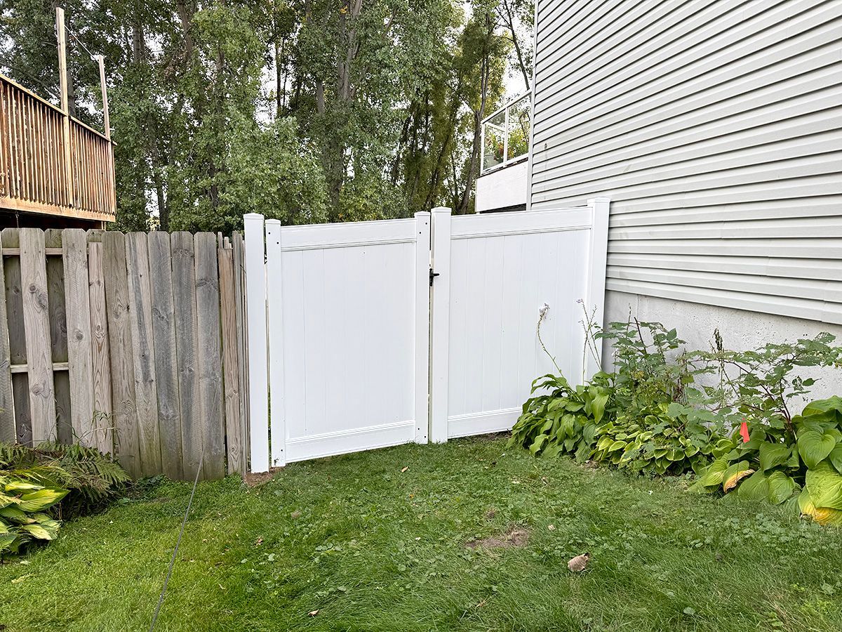 A white vinyl double gate installed between a wood fence and the side of a grey house, with green grass and foliage.