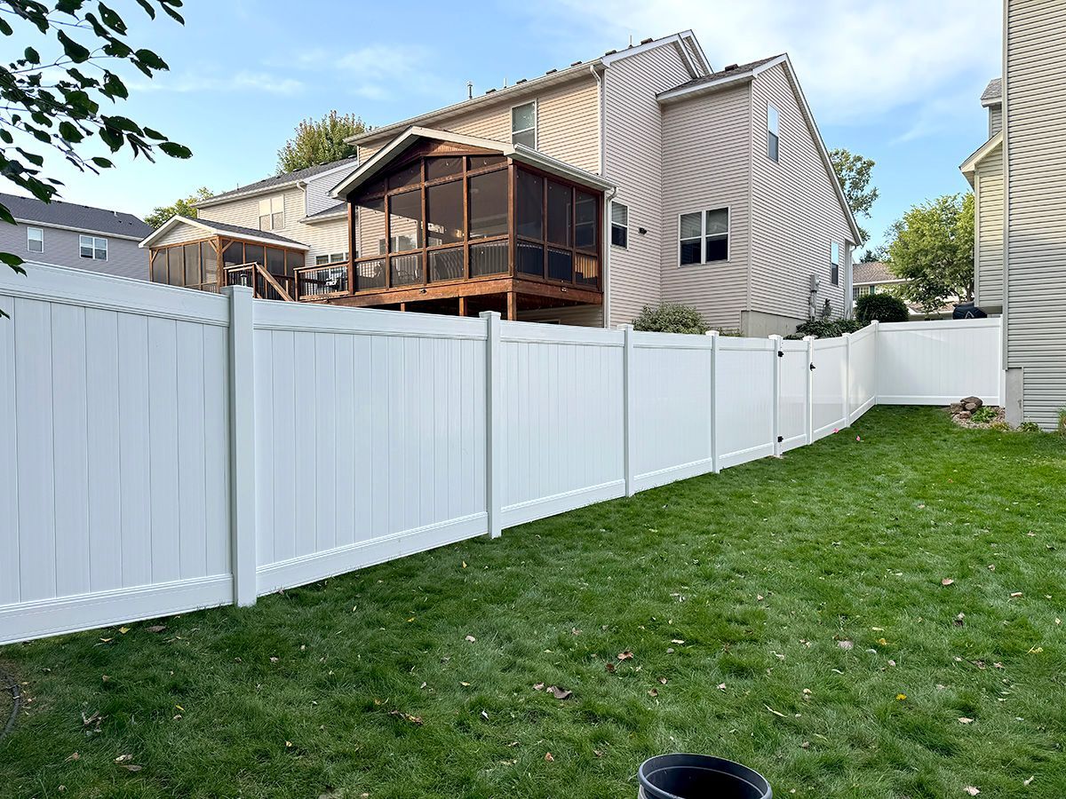 A white vinyl privacy fence curves through a residential backyard, stretching toward a two-story house with a deck.