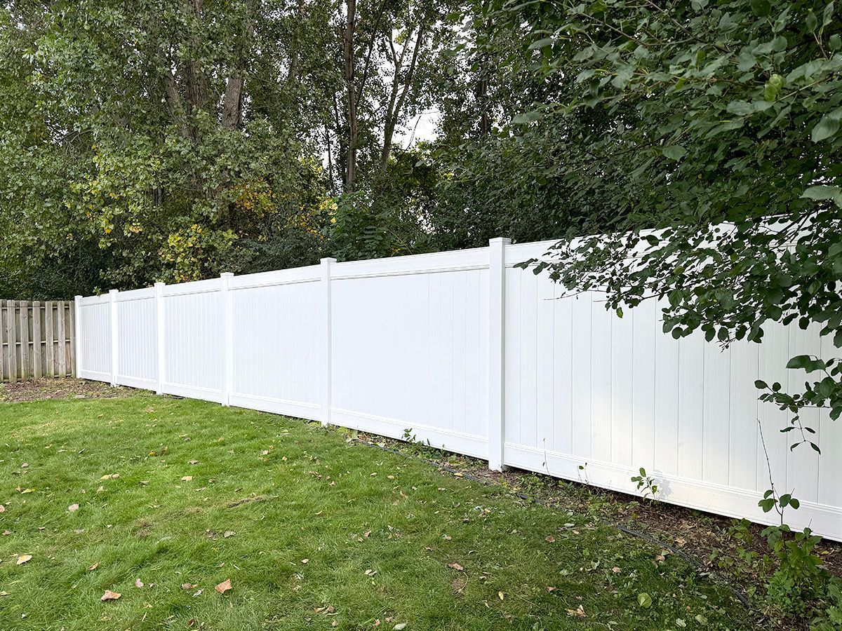 A white vinyl privacy fence lines the edge of a green lawn in front of a wooded area.