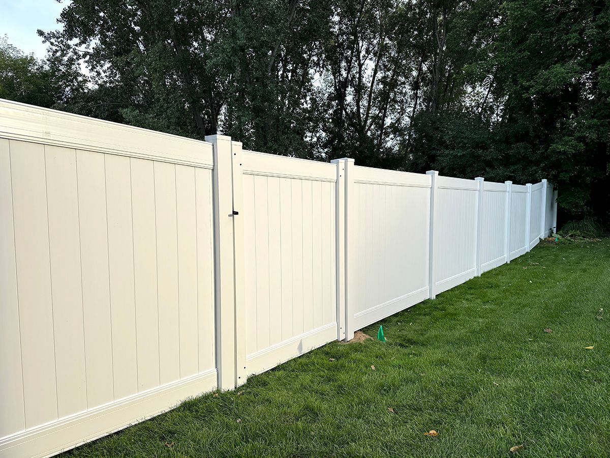 A long, white, solid-panel vinyl privacy fence stands in a grassy yard, stretching towards a background of green trees.