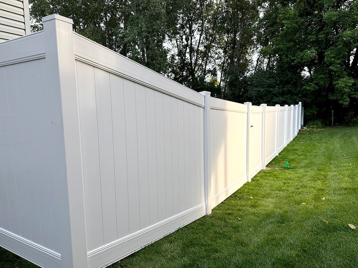 A tall, white vinyl privacy fence lines the edge of a lush green lawn, extending toward a backdrop of dense trees.