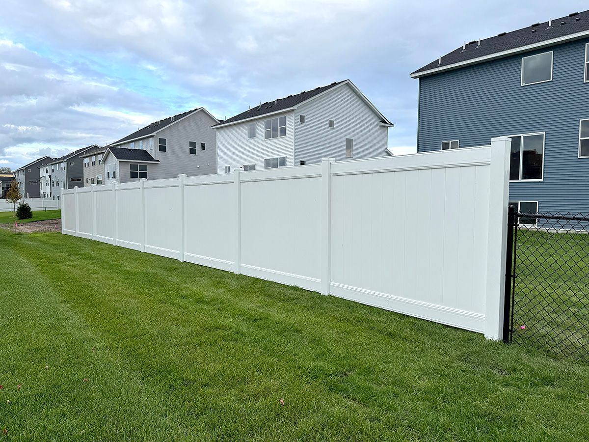 A long, white vinyl privacy fence runs across a green residential lawn under a cloudy sky, near several suburban homes.
