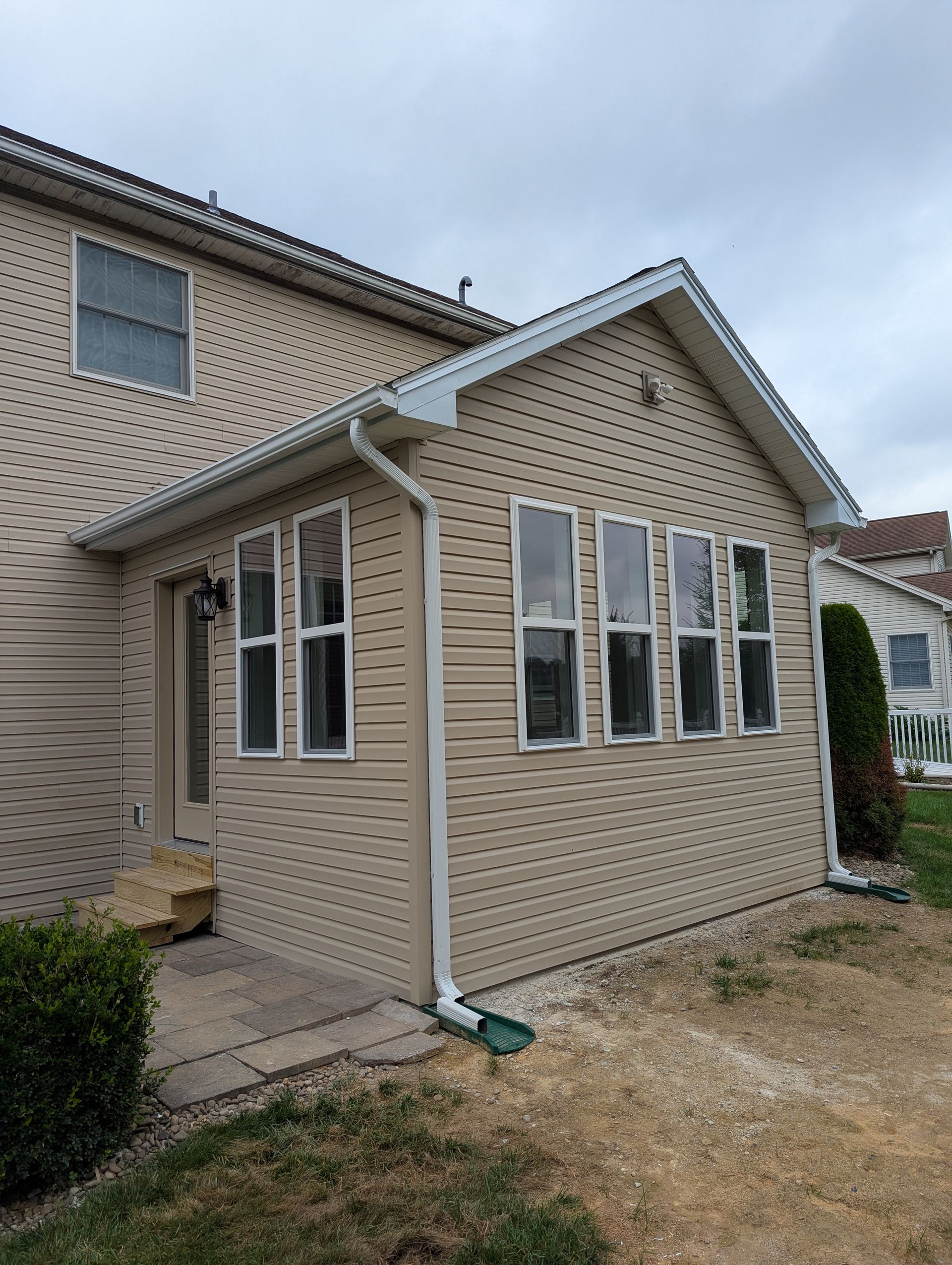 Tan siding on a house with an attached sunroom; windows, door, and a downspout are visible.