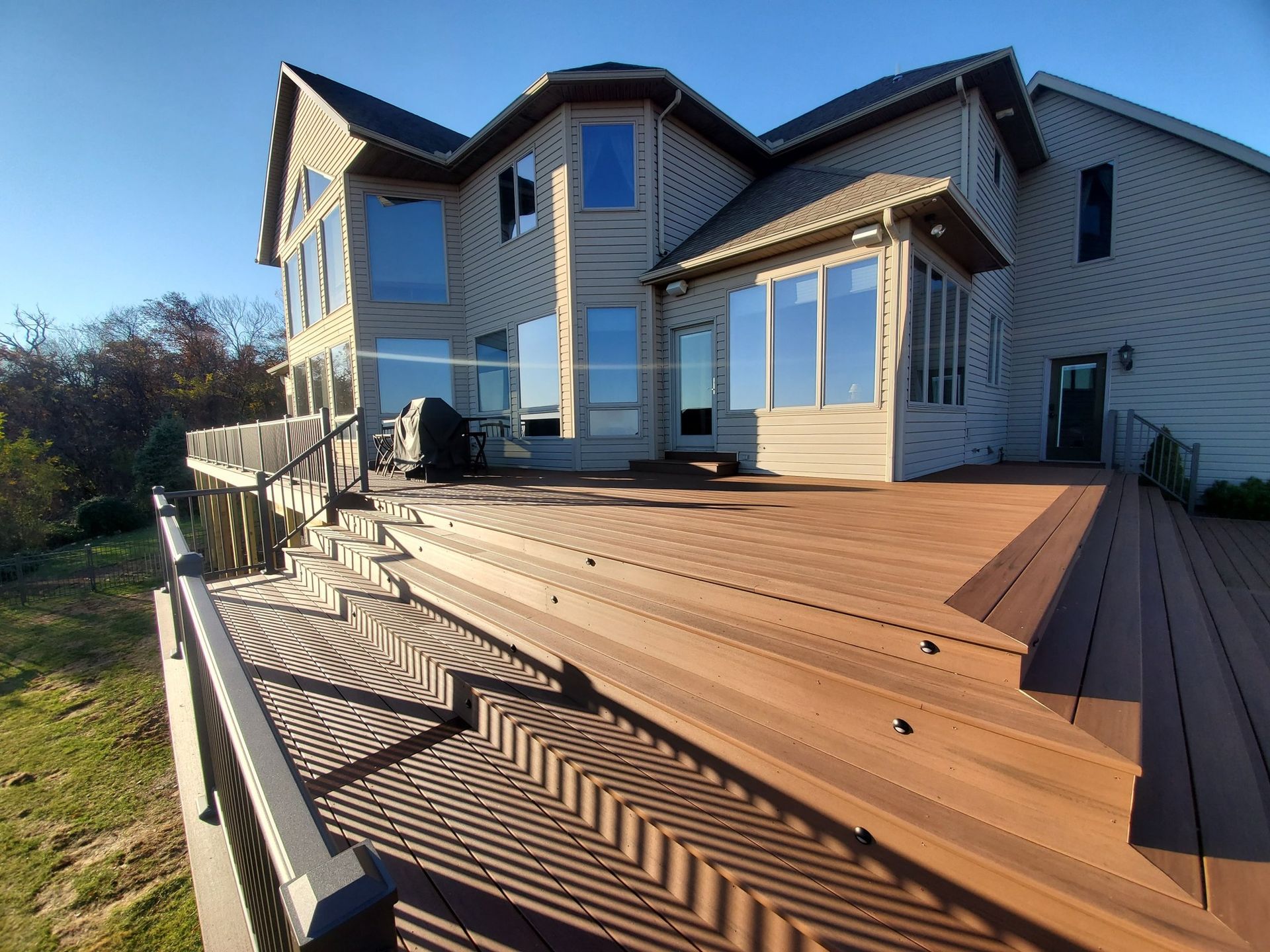 Large multi-level wooden deck attached to a two-story beige house with many windows; clear, sunny day.