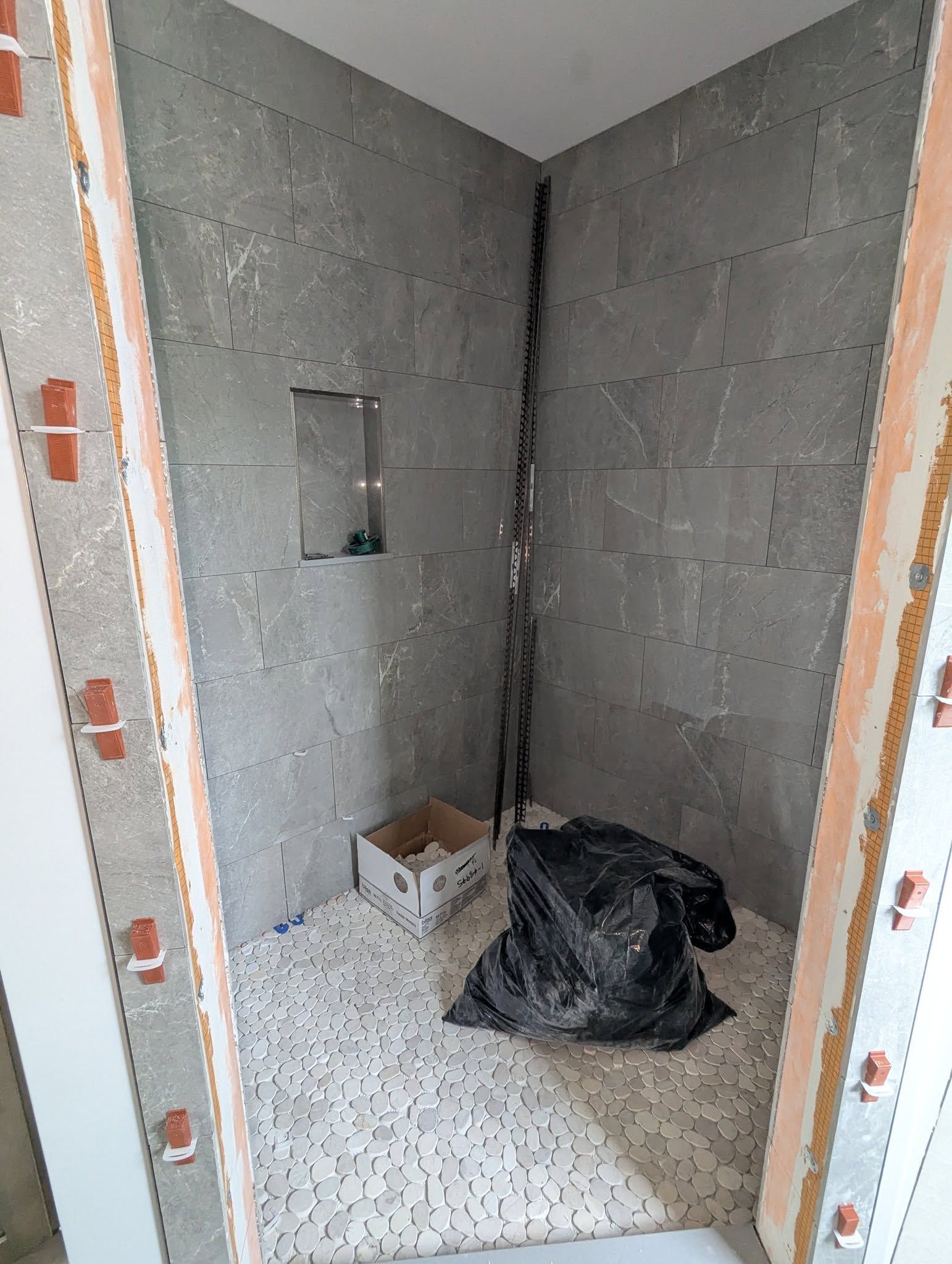 Shower stall under construction, gray tiled walls, small mosaic floor, trash bag present.