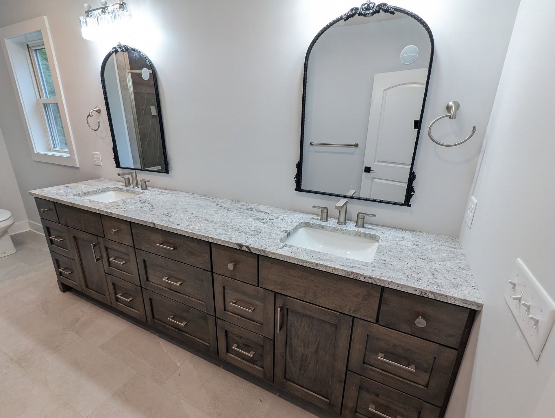 Bathroom with double vanity, dark cabinets, light granite countertop, arched mirrors, and white walls.