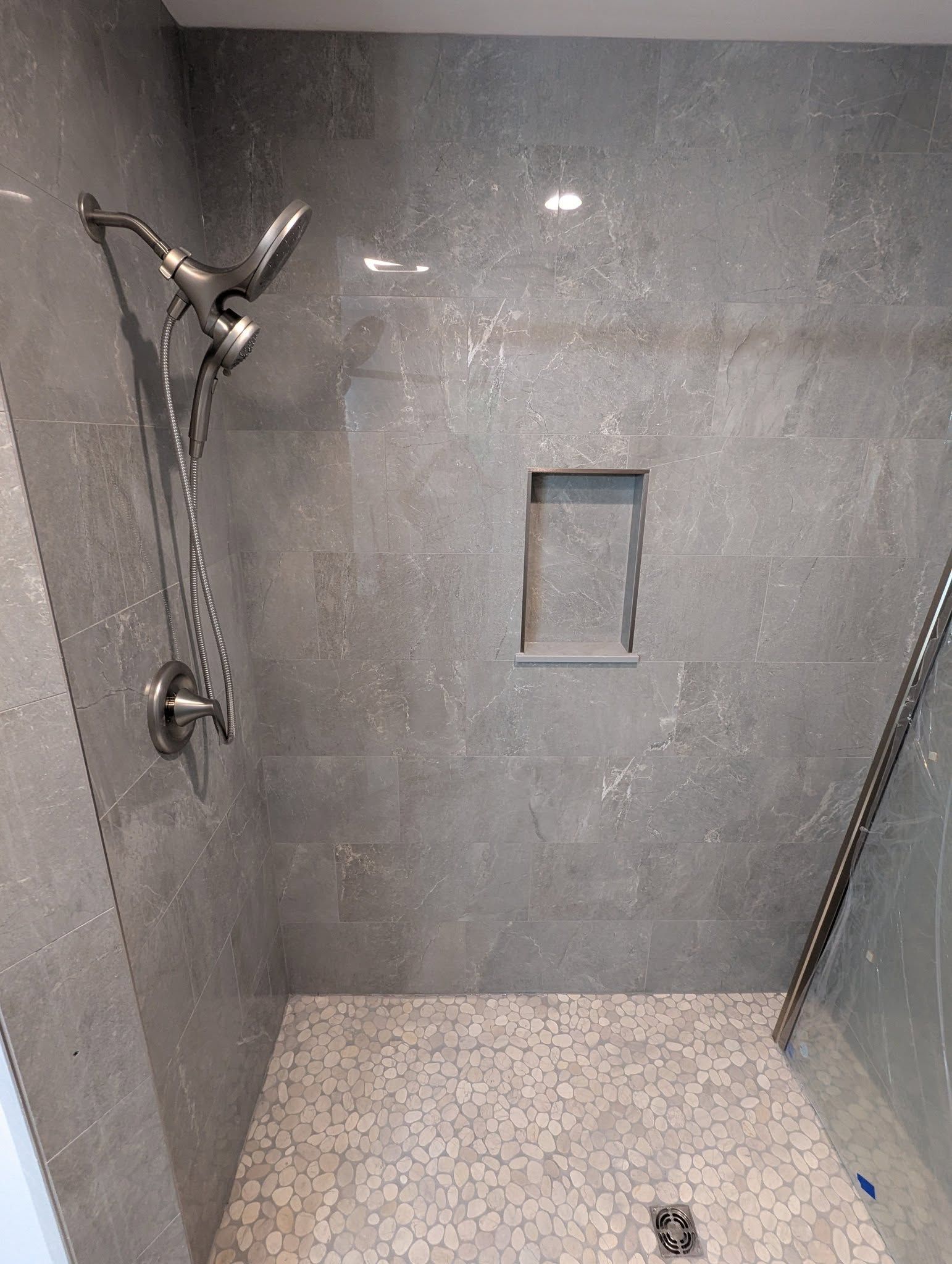Gray tiled shower with pebble floor, showerhead, and small recessed shelf.