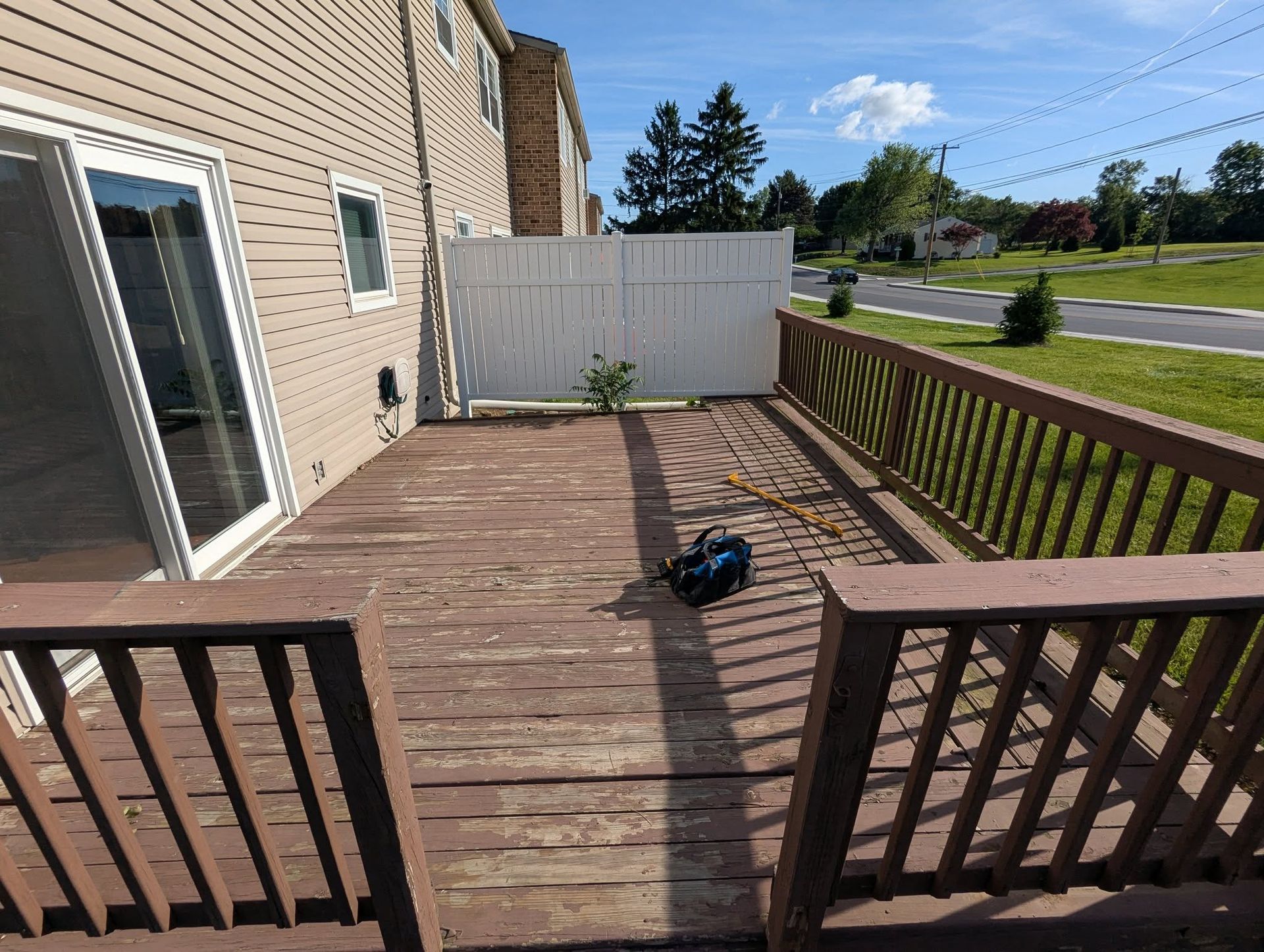 Wooden deck with brown railings, a sliding glass door, white fence, and a sunny, outdoor setting.