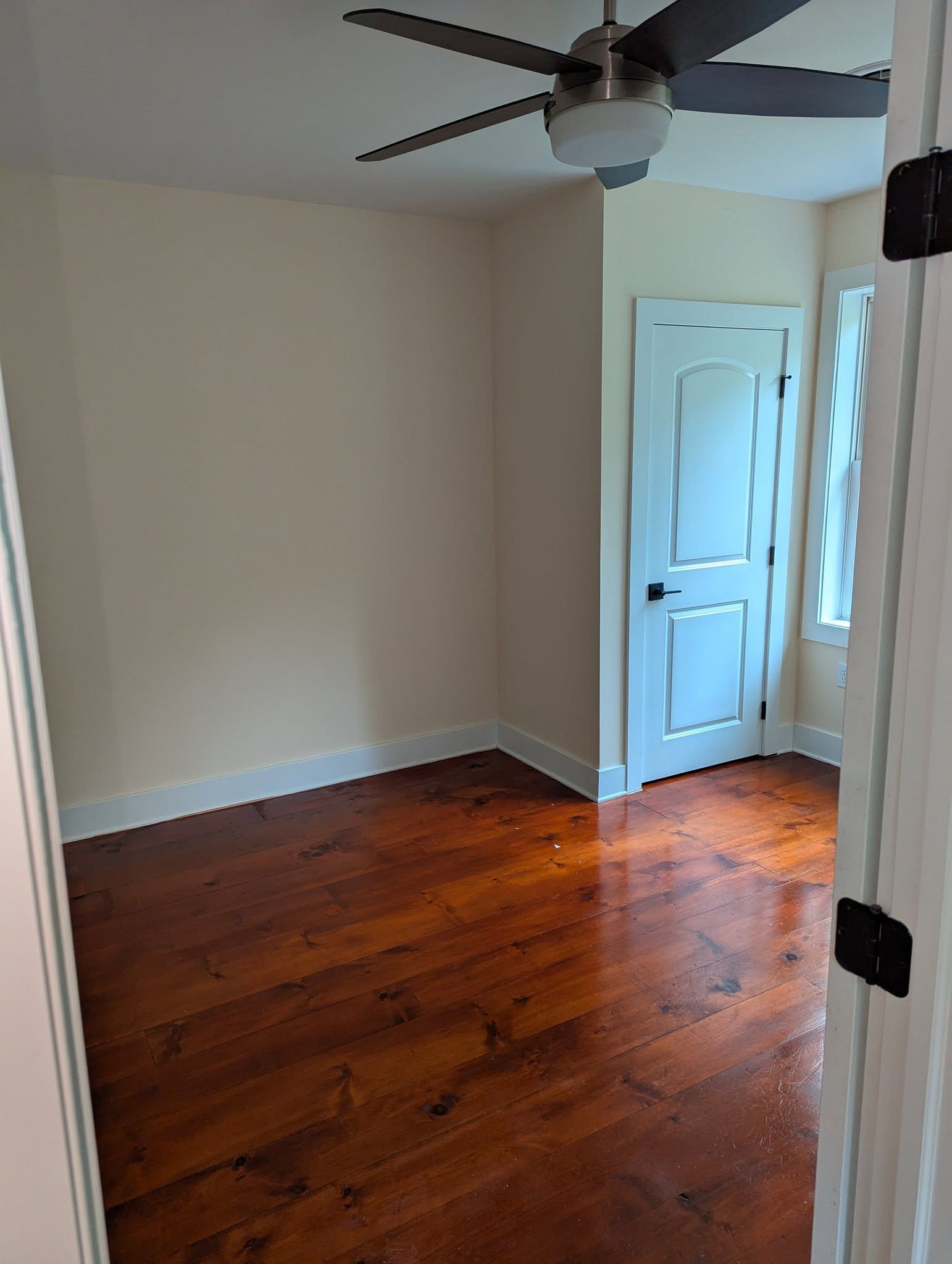Empty room with stained wood floor, white door, and beige walls. Ceiling fan and a small window are visible.