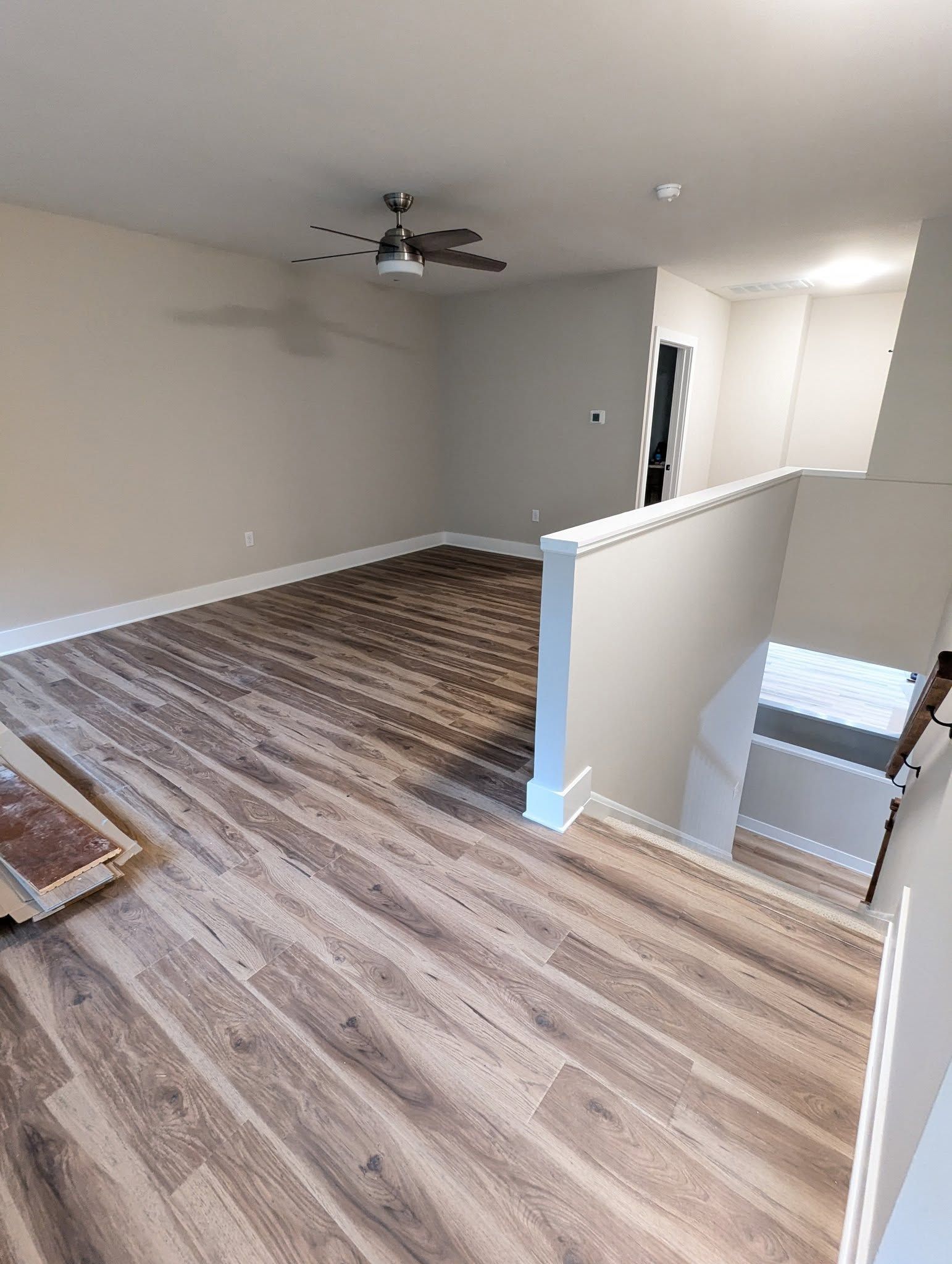Room with wood-look flooring, beige walls, and a staircase. A ceiling fan and doorway are visible.