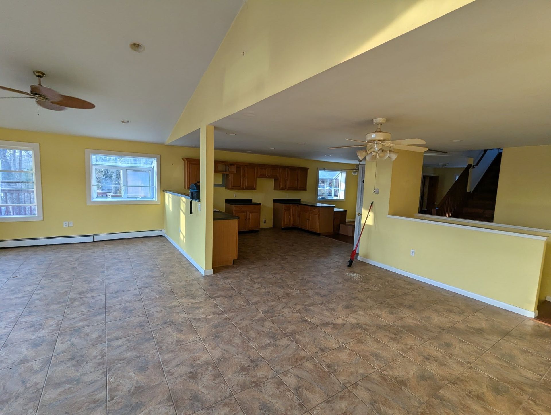 Open-concept living space with yellow walls, brown flooring, and a kitchen area. Two ceiling fans and windows are visible.