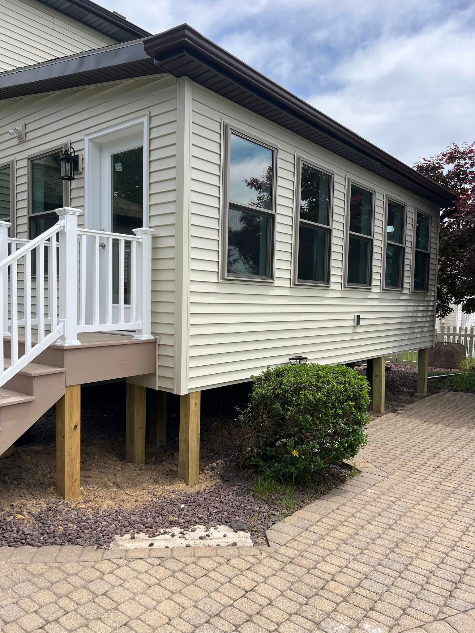 Beige-sided porch with white door and windows, supported by wooden posts. A brick pathway leads to it.