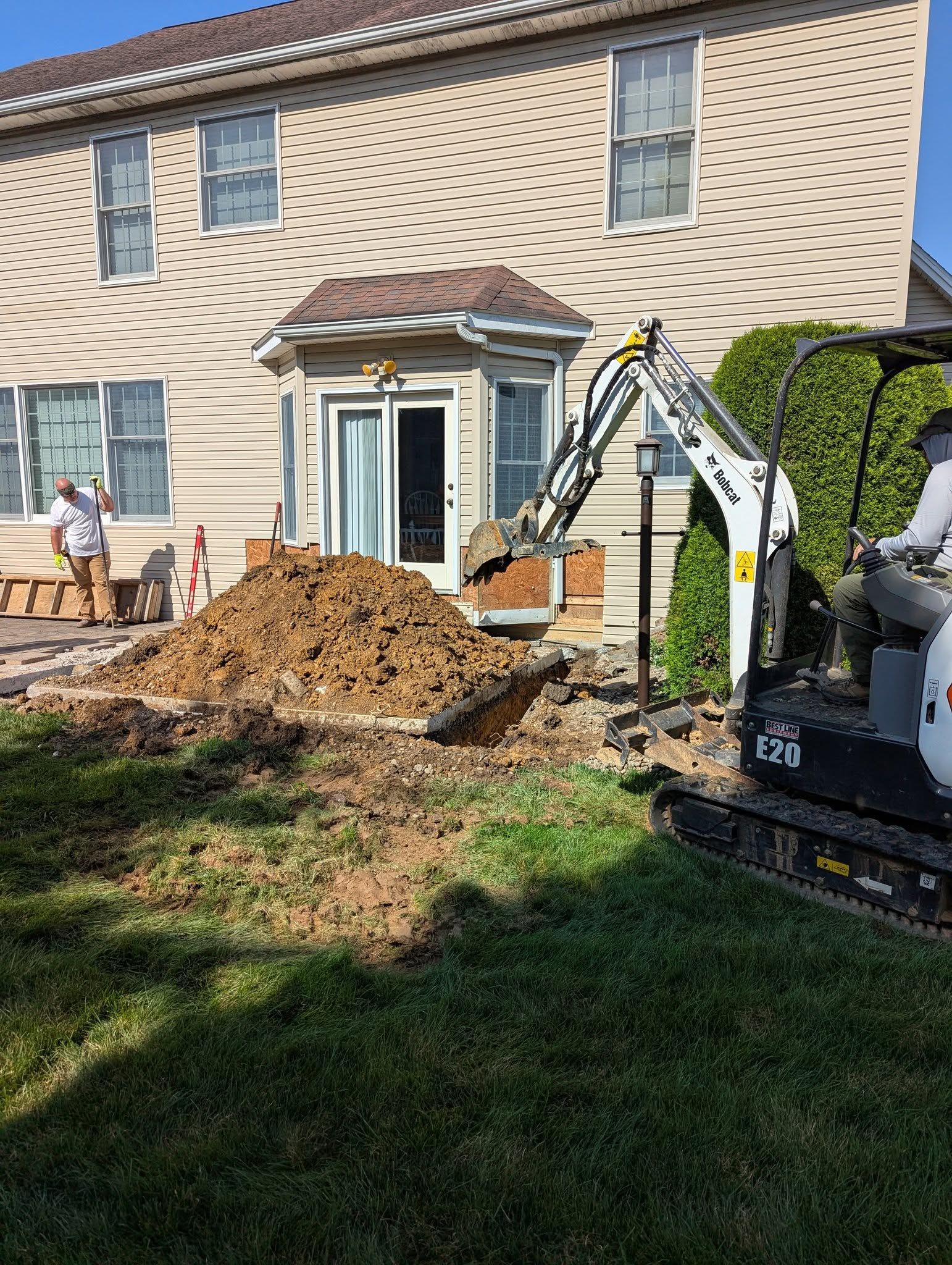 Mini excavator digging near a house, with a pile of dirt. A worker observes. Sunny day.