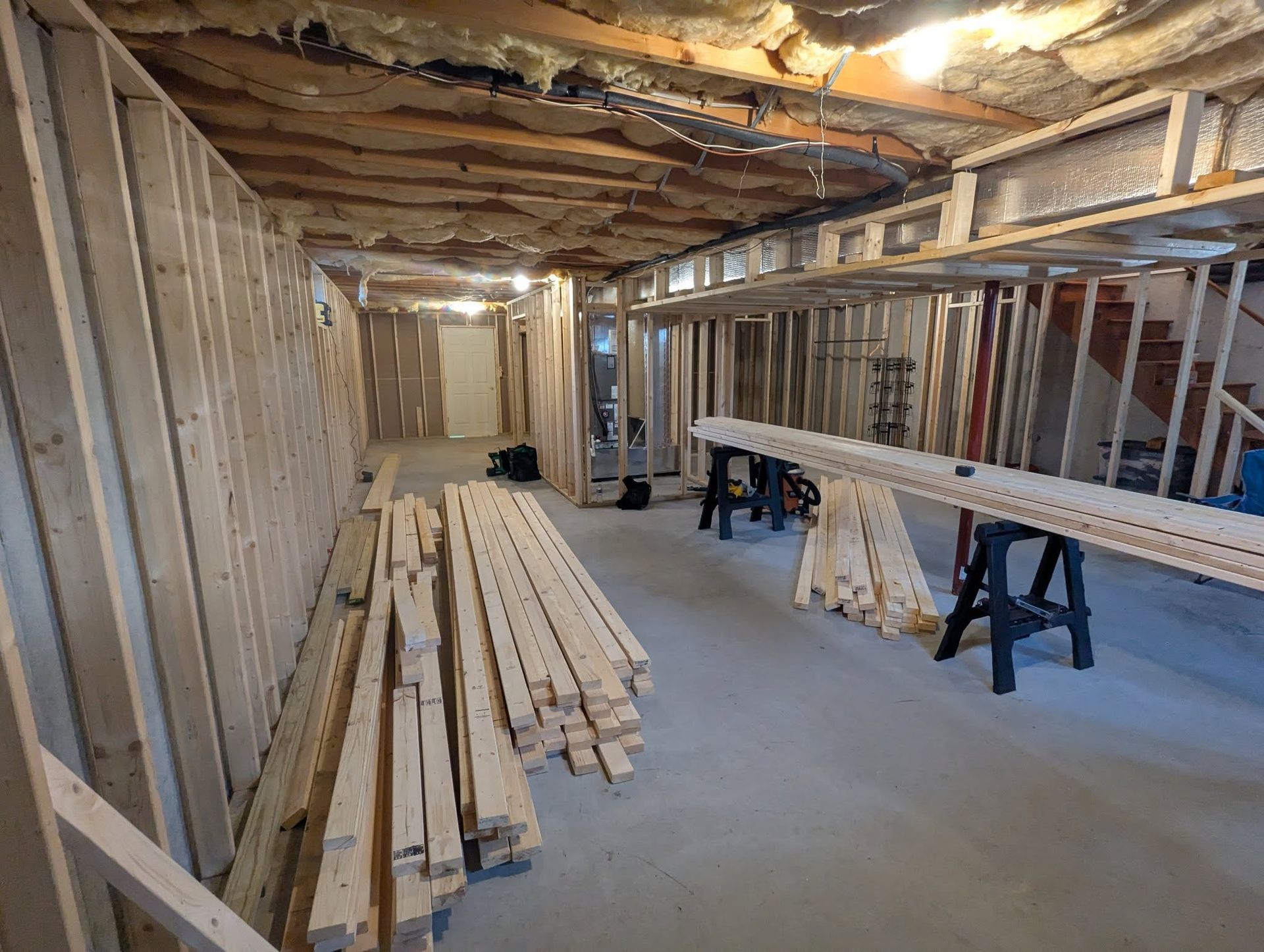 Basement under construction: wooden studs frame walls and ceiling, lumber on floor.
