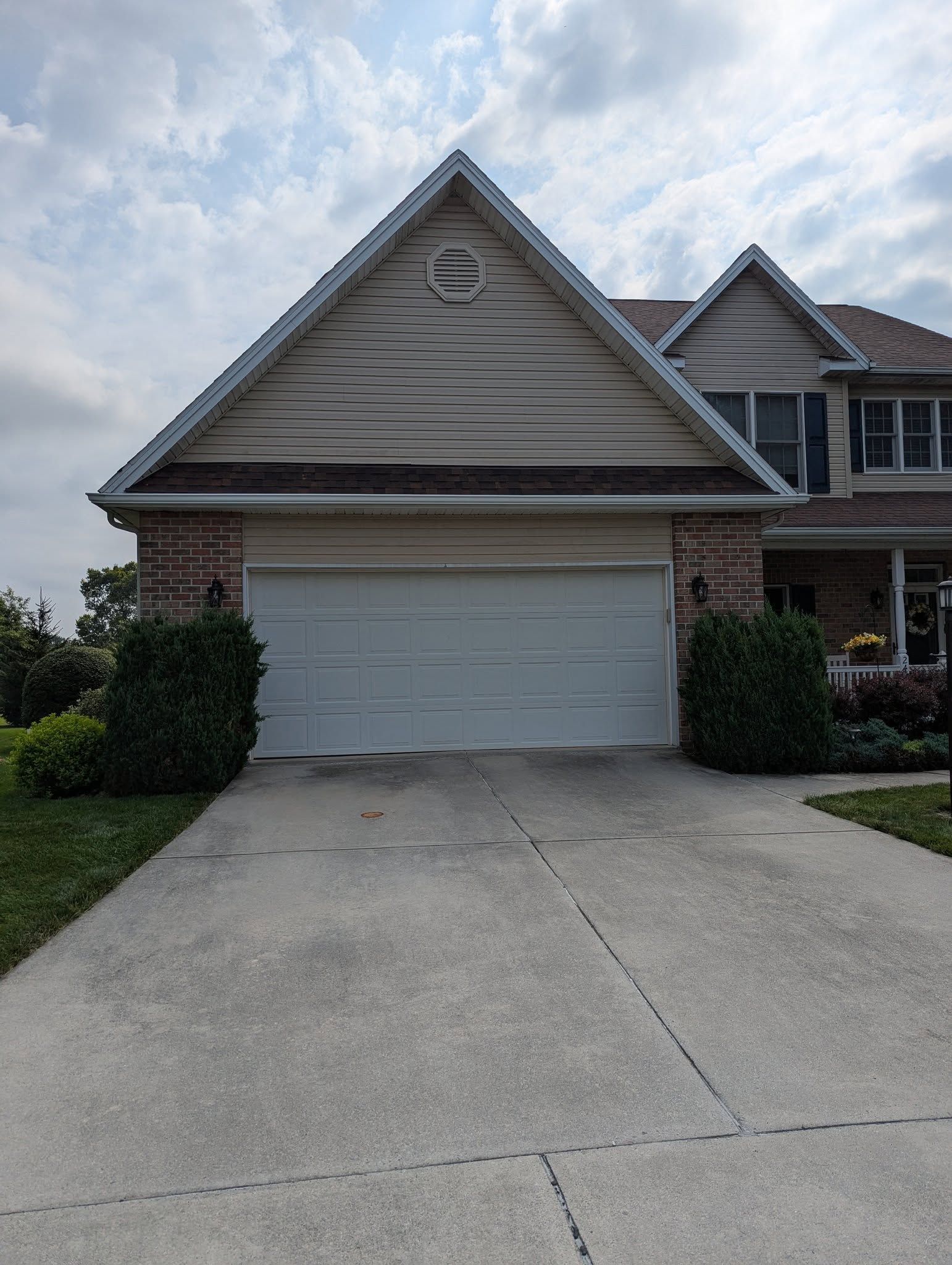 Two-car garage of a house, light-colored siding and a triangular roof. Concrete driveway with two bushes on either side.