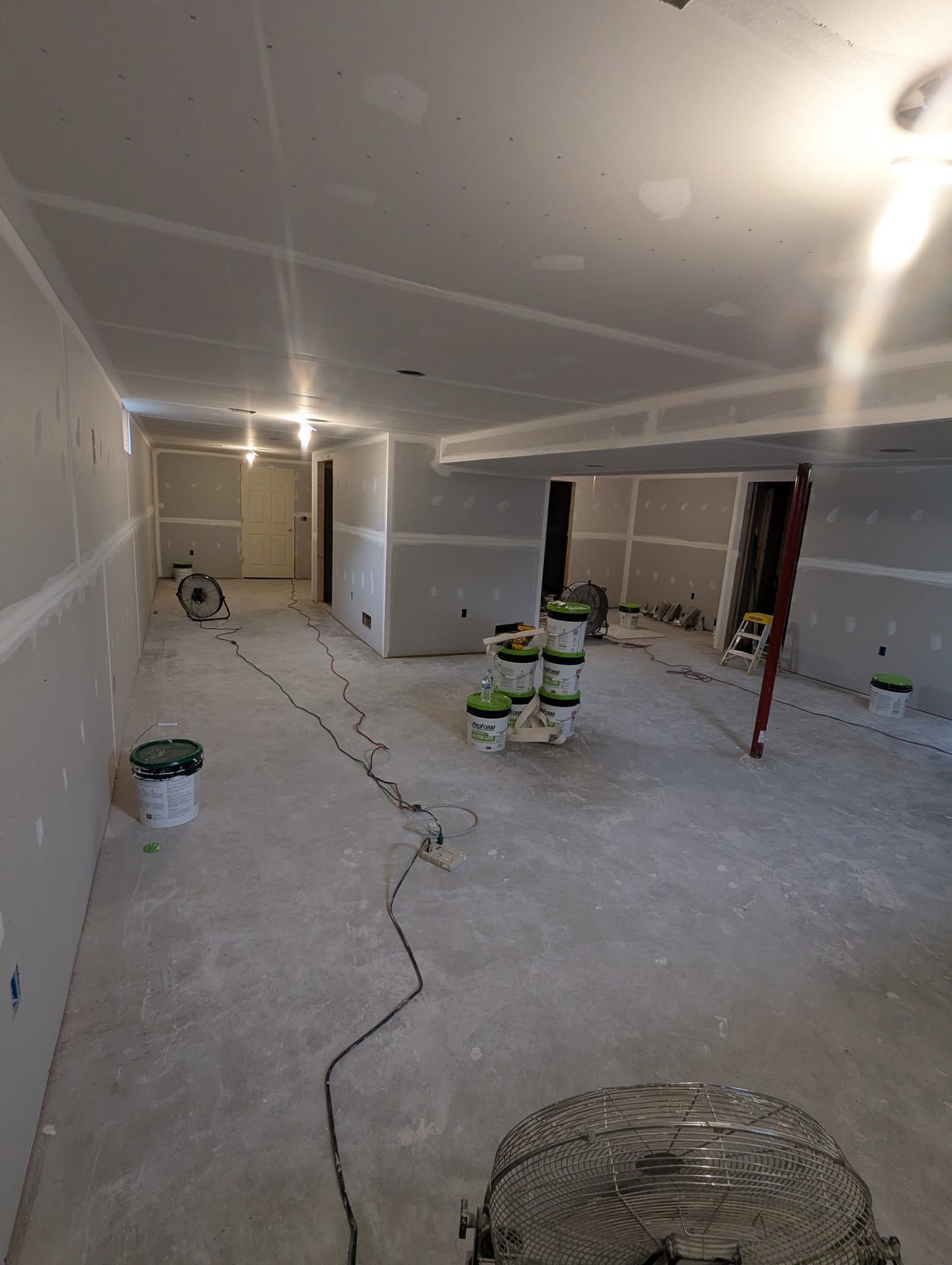 Interior of a room under construction, with drywall, paint buckets, and a fan on the floor.