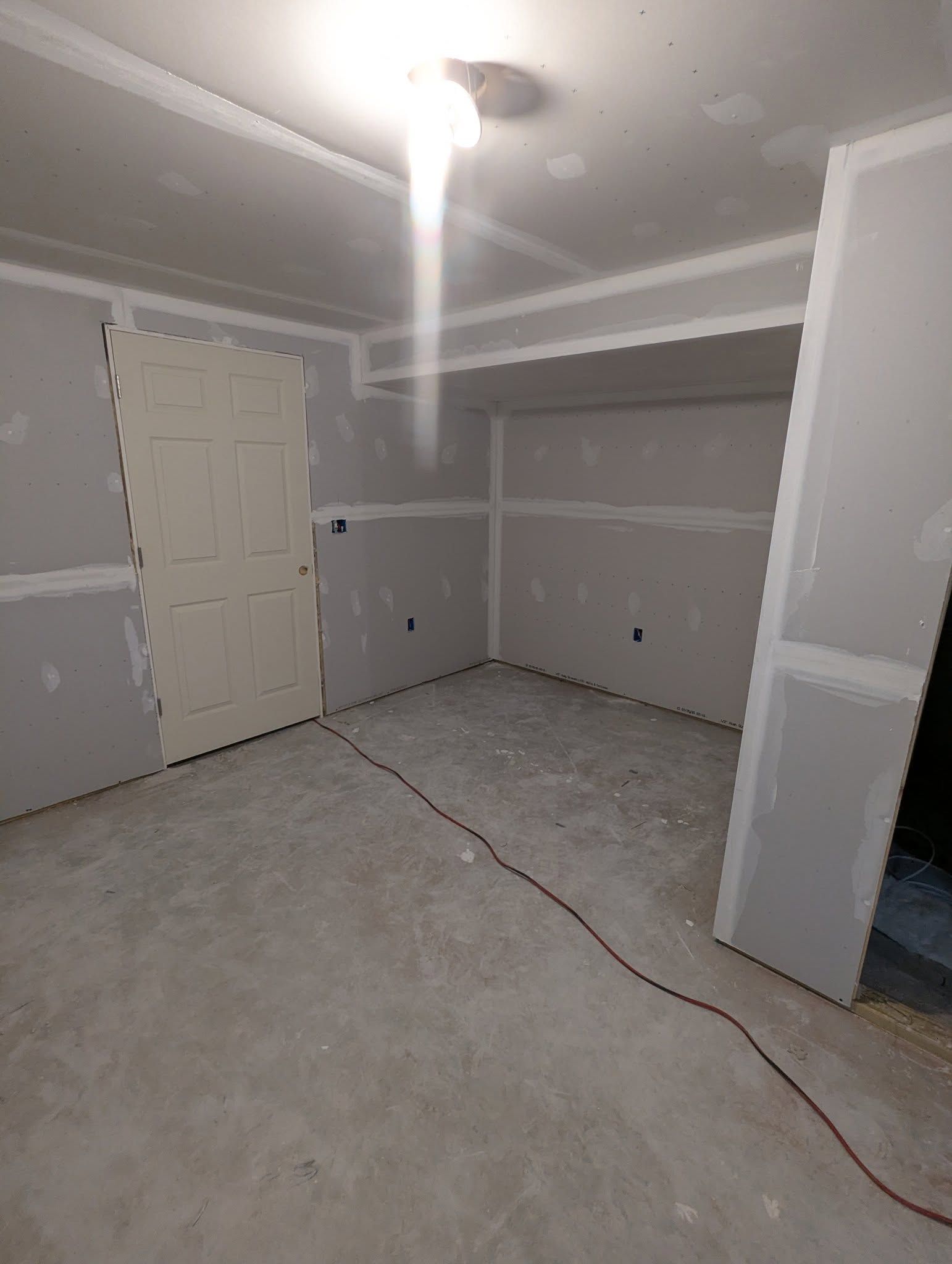 Room under construction, drywall and door installed, bare concrete floor, wire across floor.