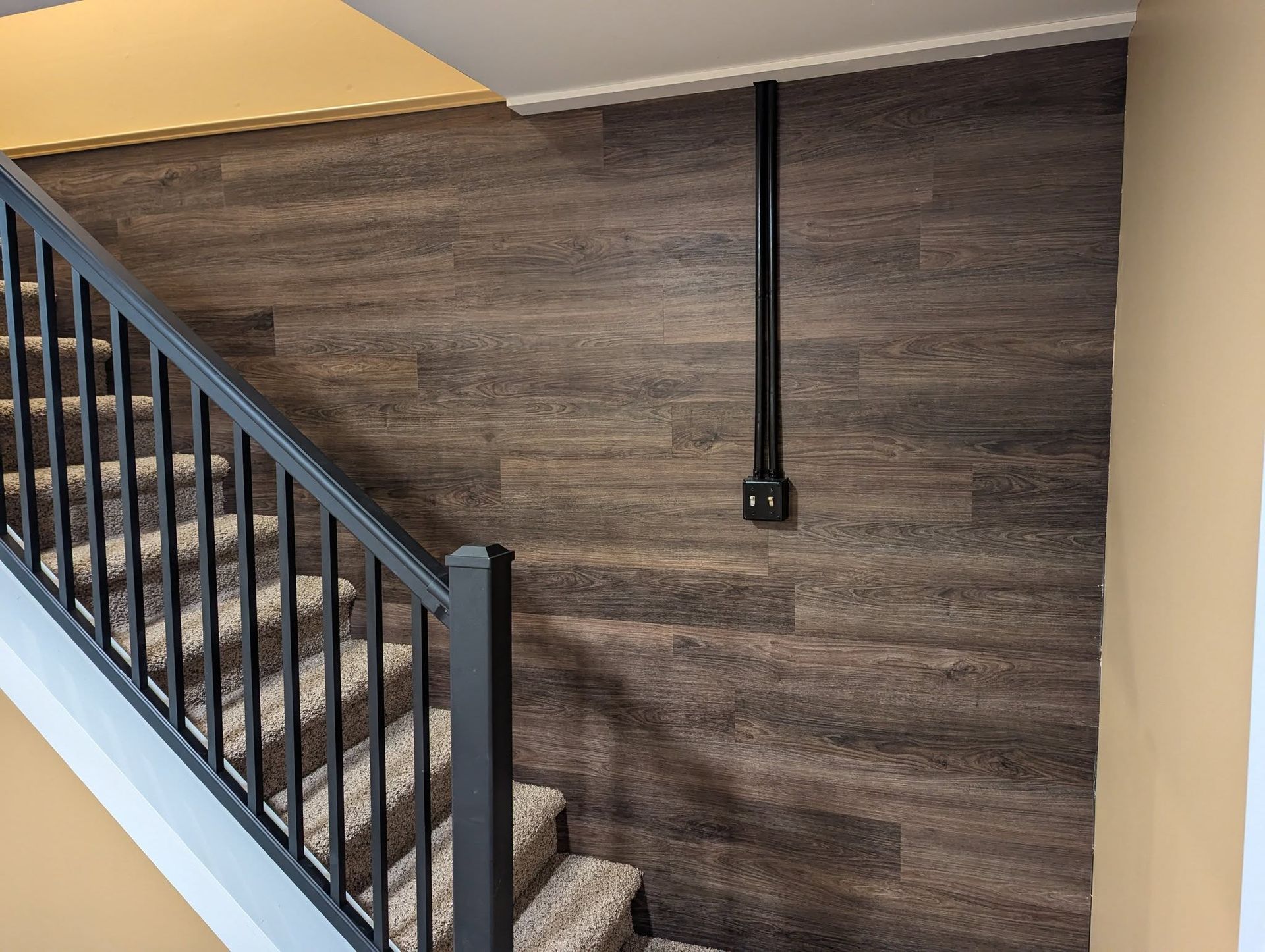Staircase with black railing and brown wood-paneled wall, black electrical conduit and outlet. Beige walls and carpeted steps.