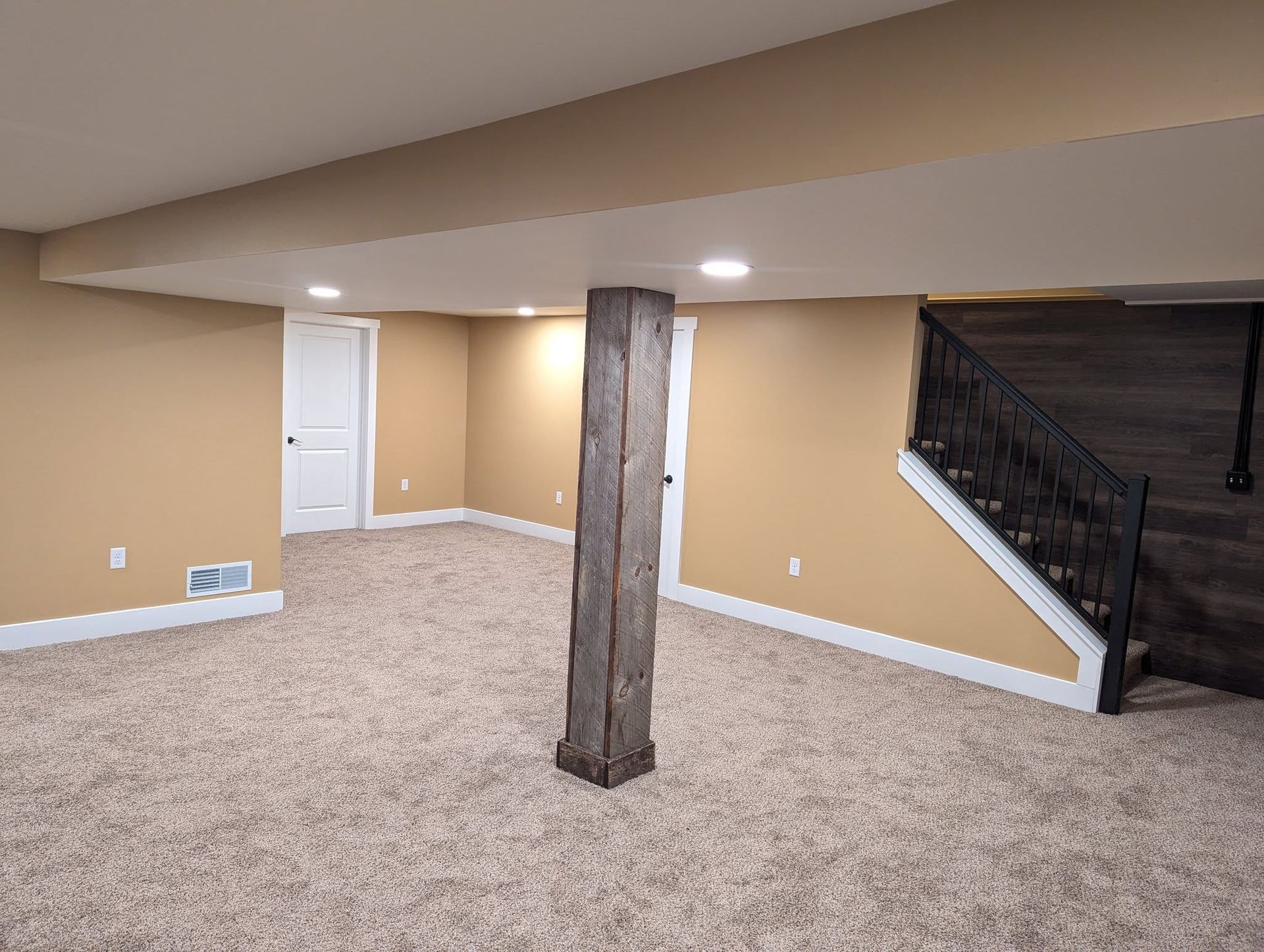 Finished basement with beige walls, carpet, and exposed support beam. White trim, door, and stairs with black railing.