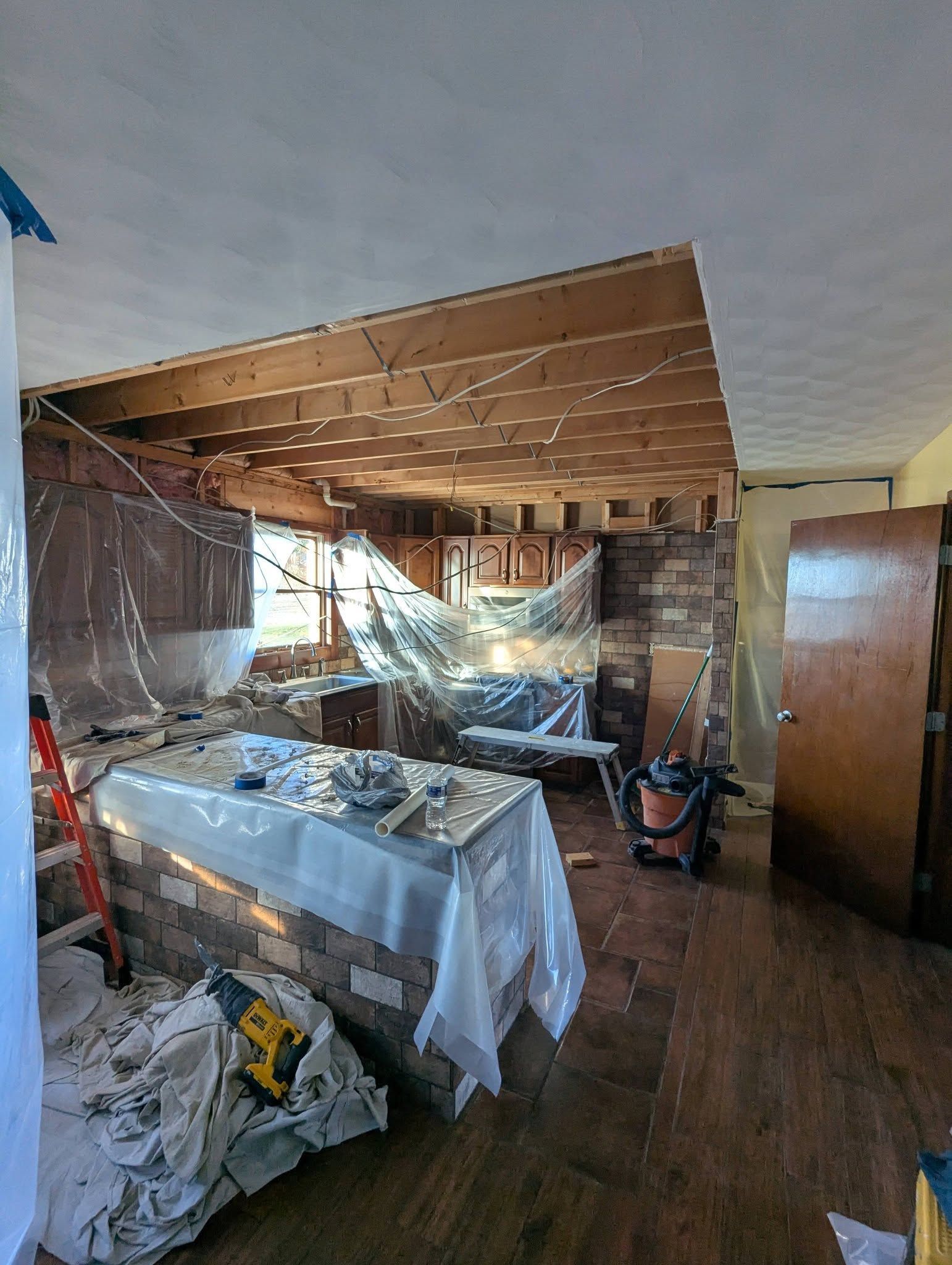 Kitchen under renovation, exposed brick, covered surfaces, wood floor, open ceiling, construction tools.