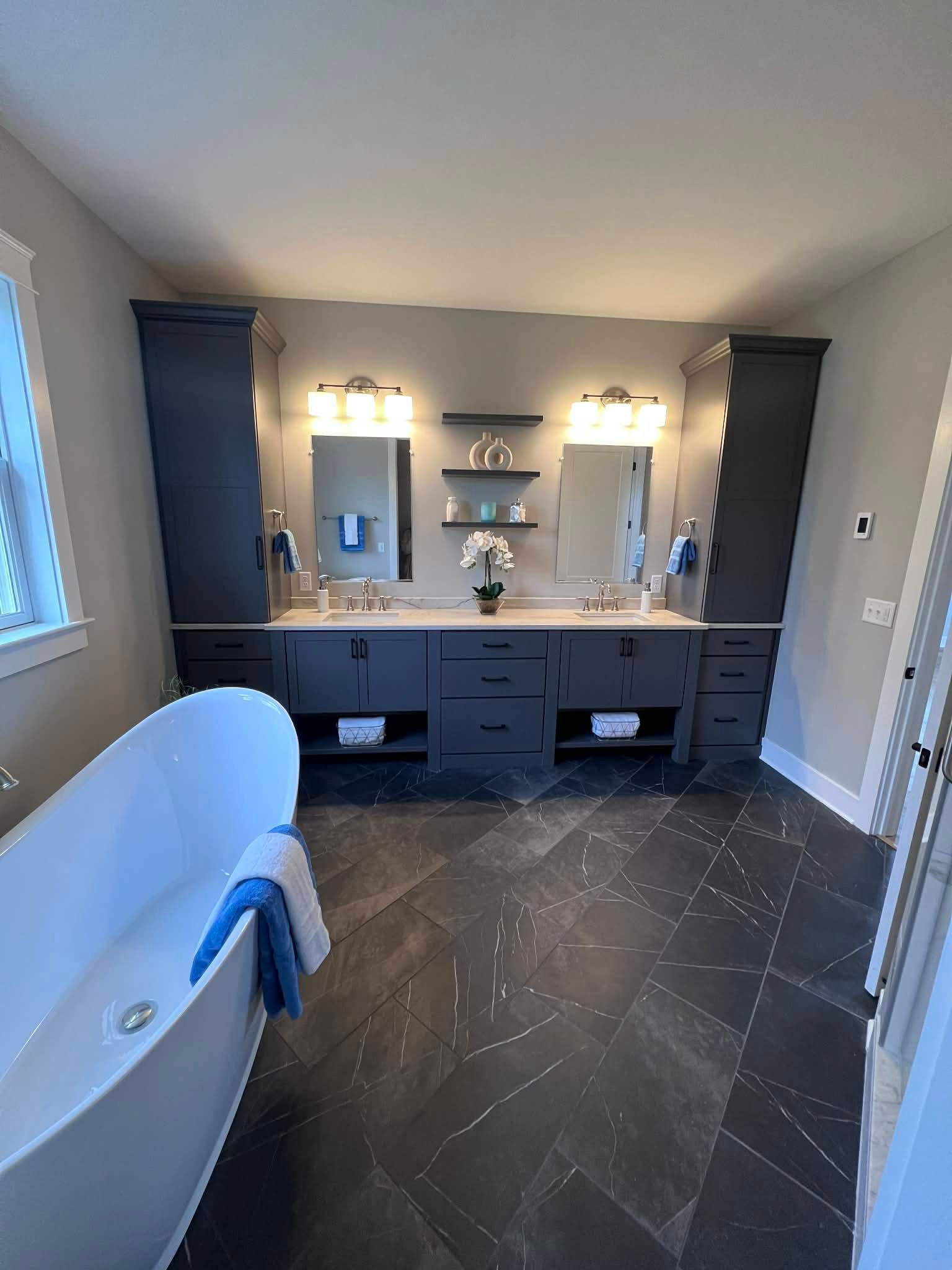Modern bathroom with a dark gray vanity, two mirrors, a freestanding tub, and dark herringbone tile.